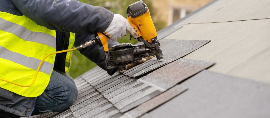 Unrecognizable roofer worker in special protective work wear and gloves, using air or pneumatic nail gun and installing asphalt or bitumen shingle on top of the new roof under construction residential building