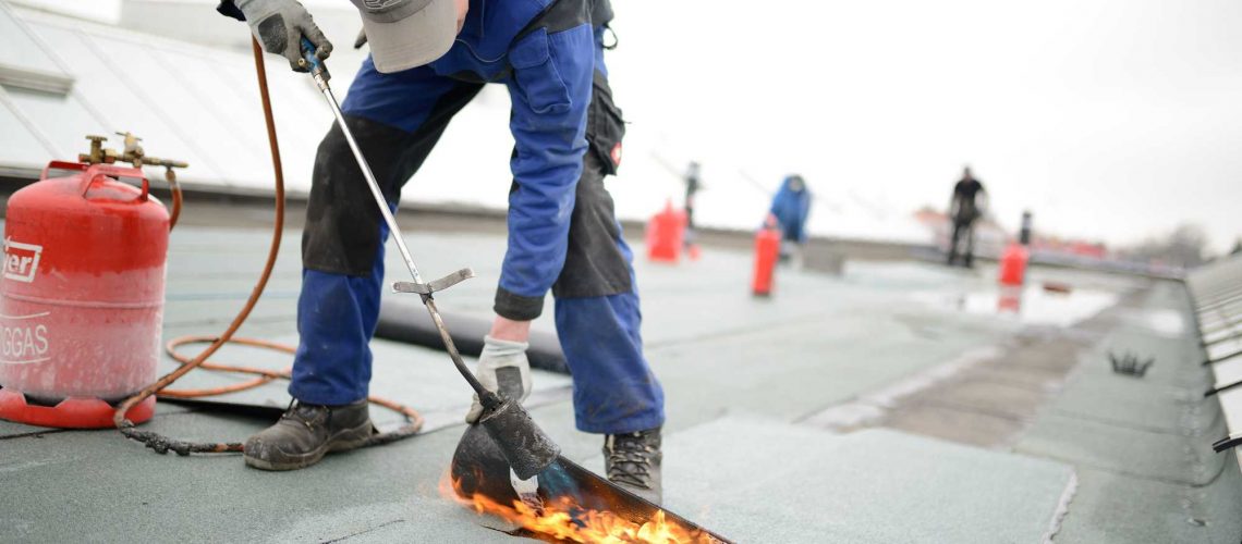 Flachdach Reparatur: Dachdecker verschweißt Dachabdichtung. Dachsanierung mit Gasbrenner. Immer Feuerlöscher vorhalten.